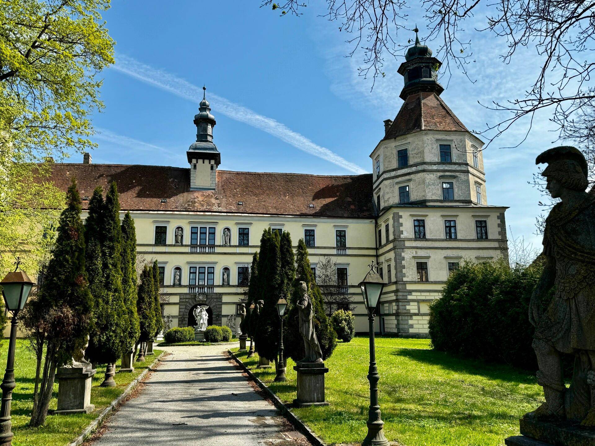 Historisches Schloss mit hohen Bäumen und einem gepflegten Rasen, der zum Eingangsbereich führt.