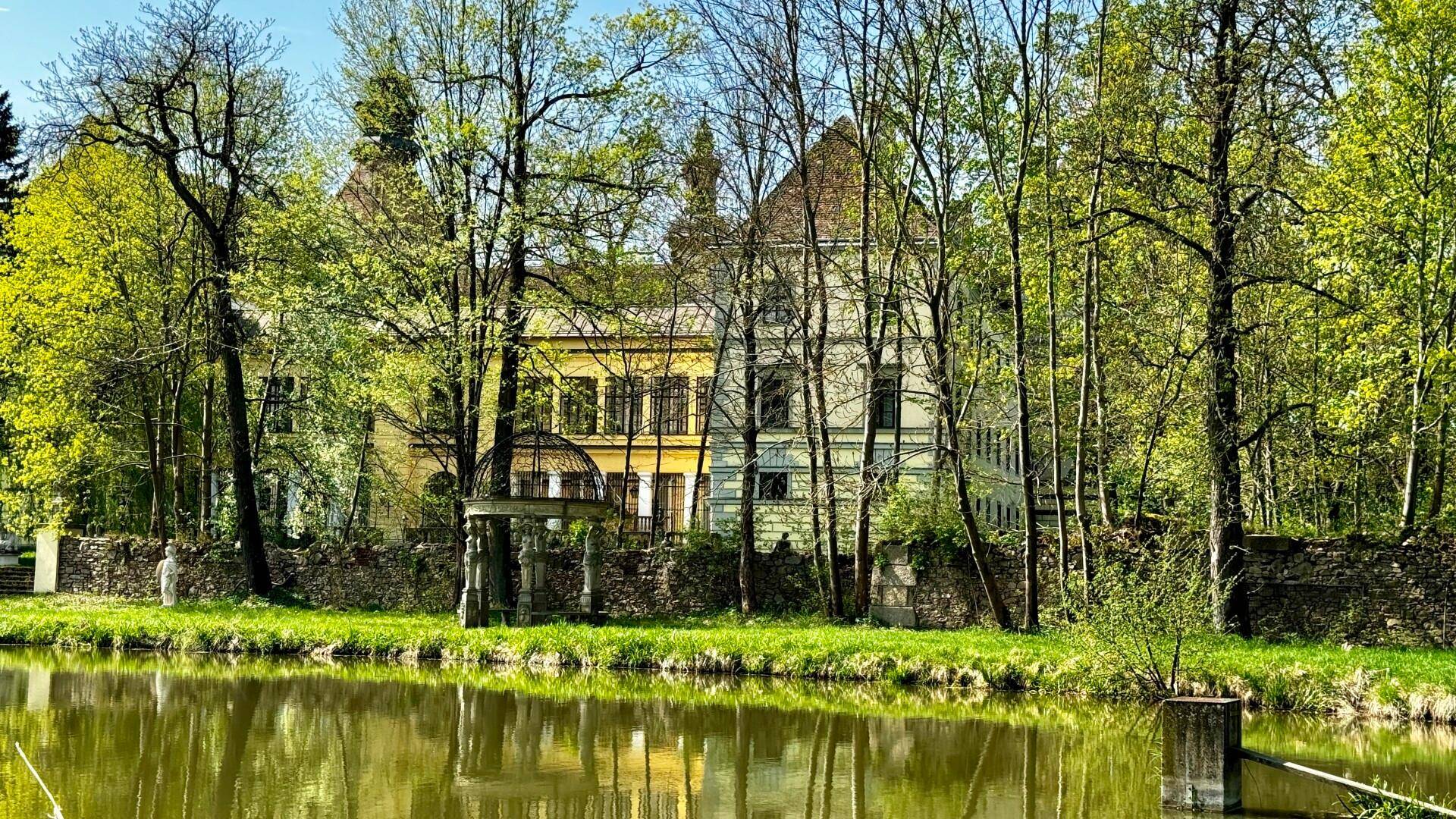 Schlossgebäude umgeben von Bäumen, gespiegelt in einem ruhigen Teich, der eine idyllische Landschaft schafft.