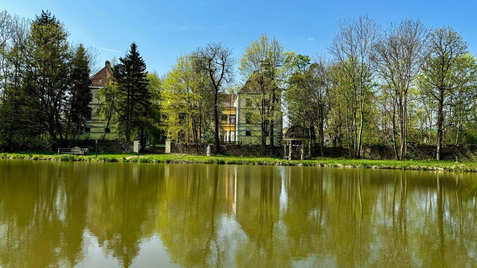 Weitläufige Ansicht des Schlosses über einen Teich, umgeben von üppiger Vegetation und altem Baumbestand.