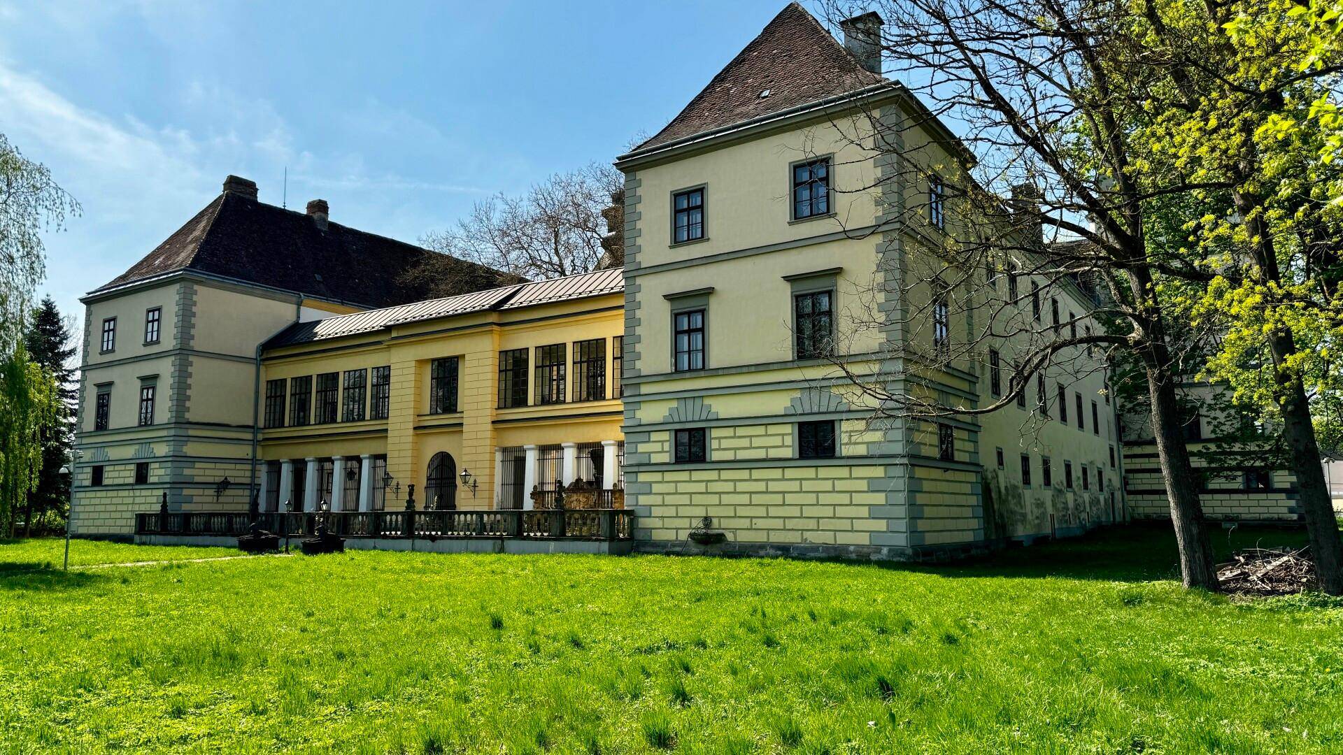 Seitenansicht des historischen Schlosses mit einem weitläufigen grünen Rasen und hohen Bäumen im Hintergrund.