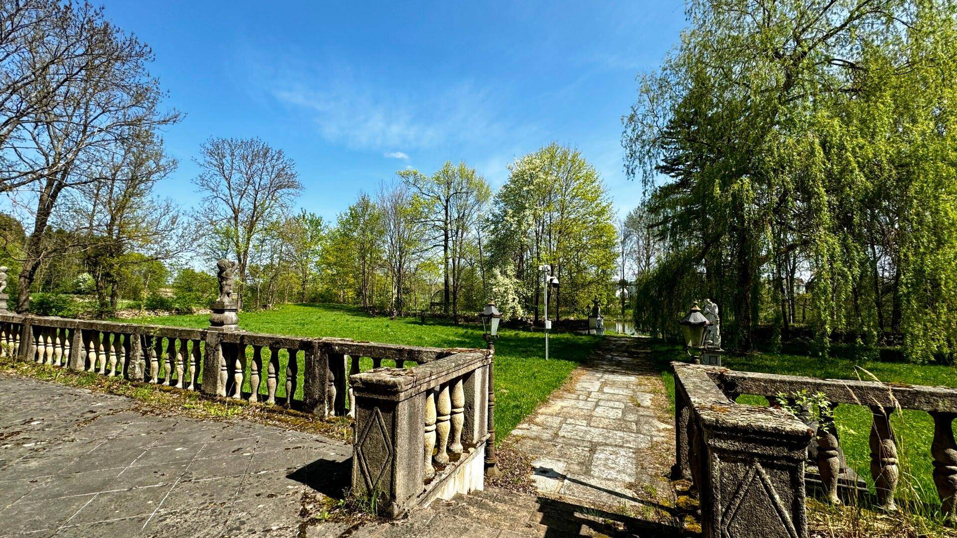 Historische Steinbalustrade und ein gepflasterter Weg, der zu einem weitläufigen grünen Parkbereich führt.