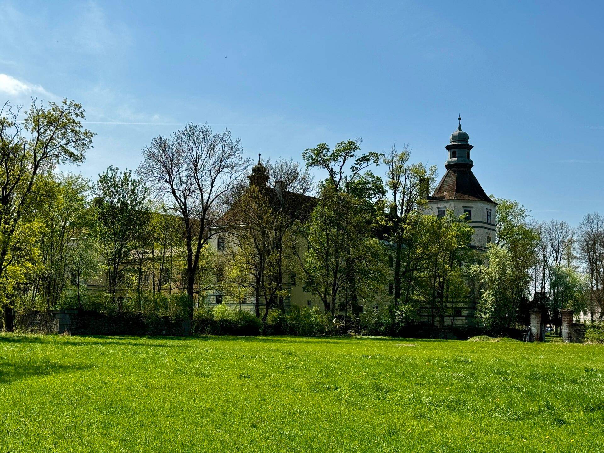 Das Schloss ist teilweise von Bäumen verdeckt, mit einem weiten grünen Feld im Vordergrund unter blauem Himmel.