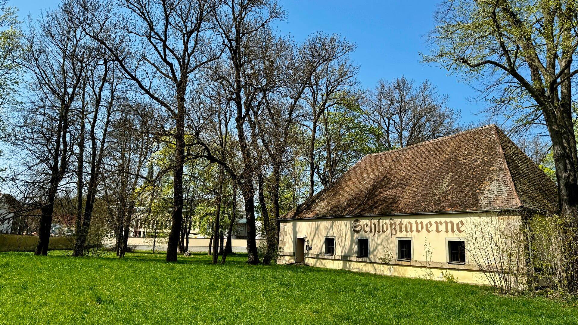 Ein charmantes Nebengebäude mit der Aufschrift "Schloßtaverne", umgeben von hohen Bäumen und grünem Rasen.