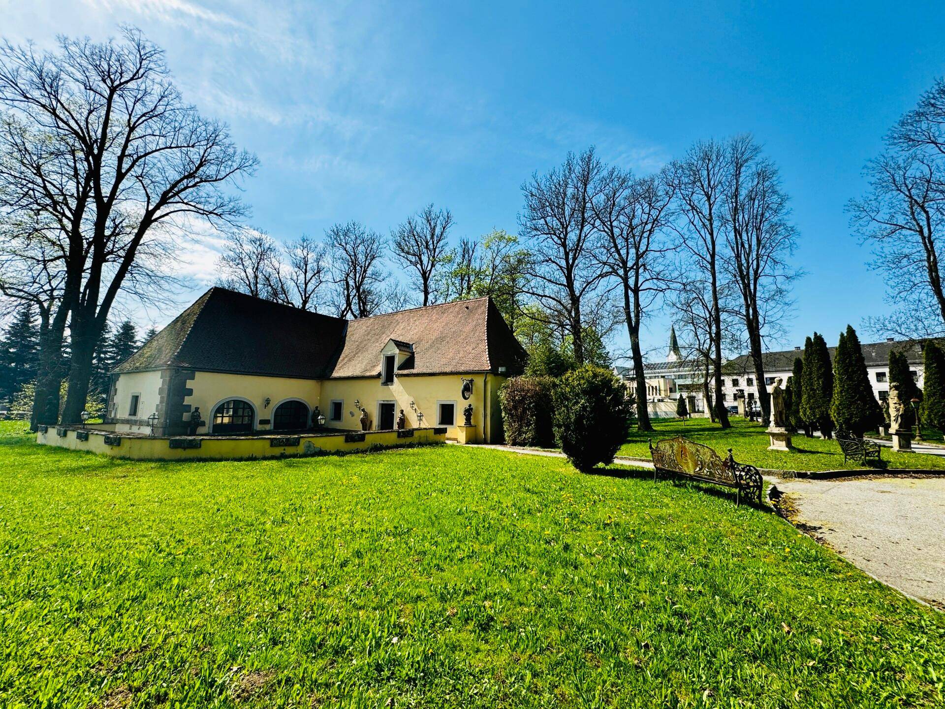 Ein kleineres Gebäude mit Satteldach auf einem weitläufigen Rasen, mit dem Hauptschloss im Hintergrund.