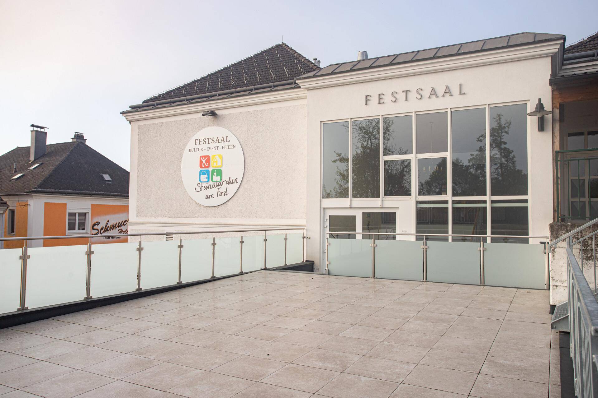 Große Außenterrasse mit Glasgeländer vor einem Gebäude mit der Aufschrift "Festsaal", modern gestaltet.