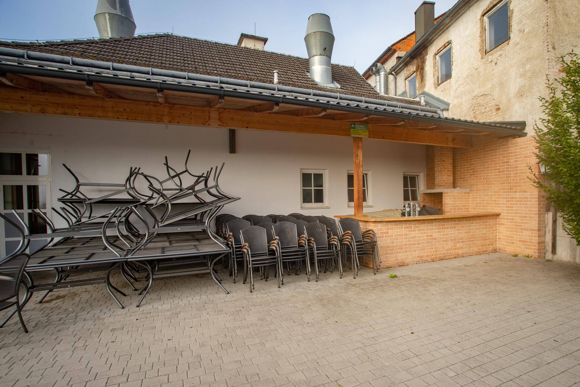 Überdachter Außenbereich mit gestapelten Stühlen und Tischen, sowie einem gemauerten Grillbereich.