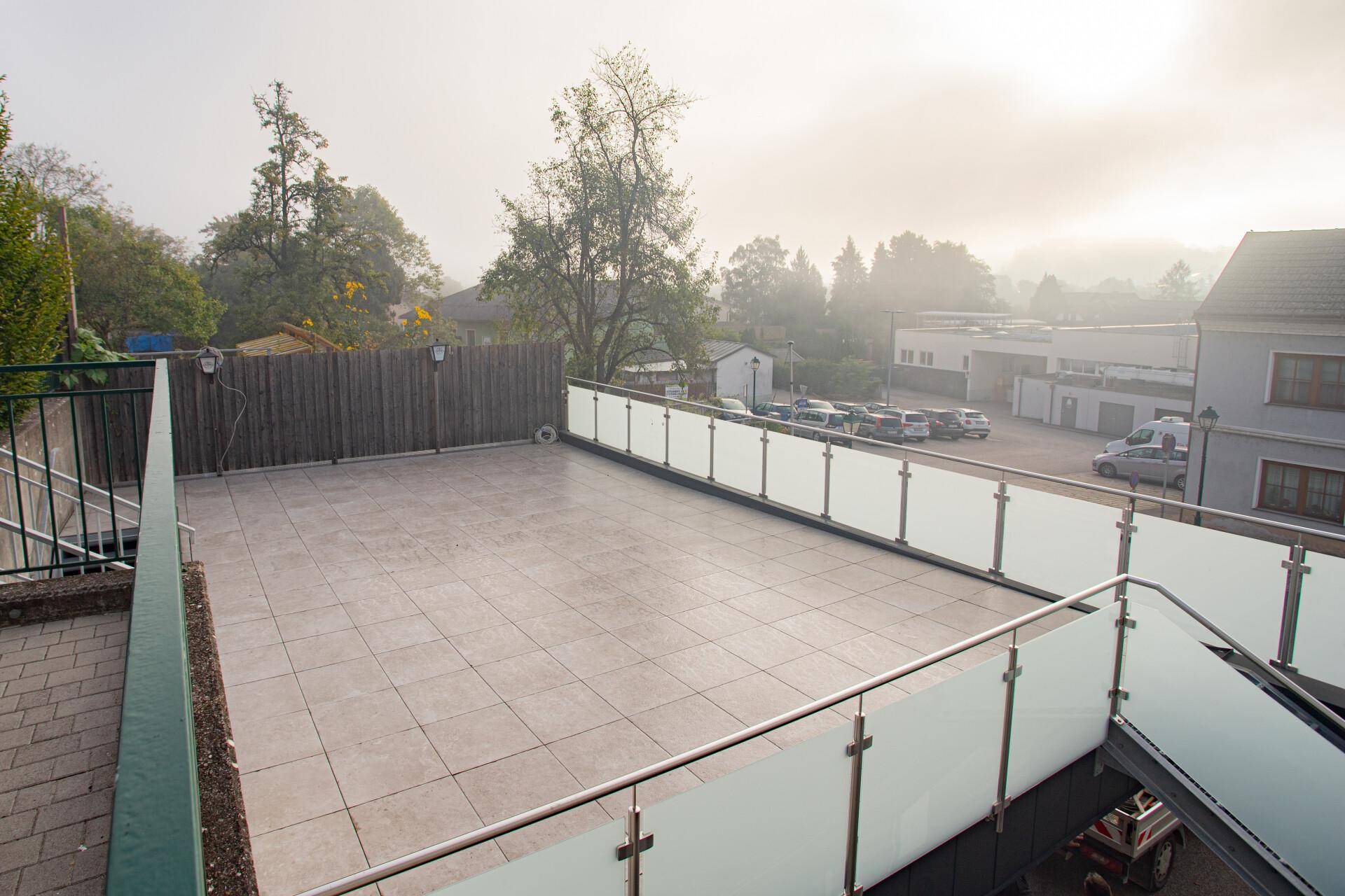 Weitläufige Terrasse mit Glasgeländer und Blick auf die umliegende Landschaft und Gebäude, sehr offen.