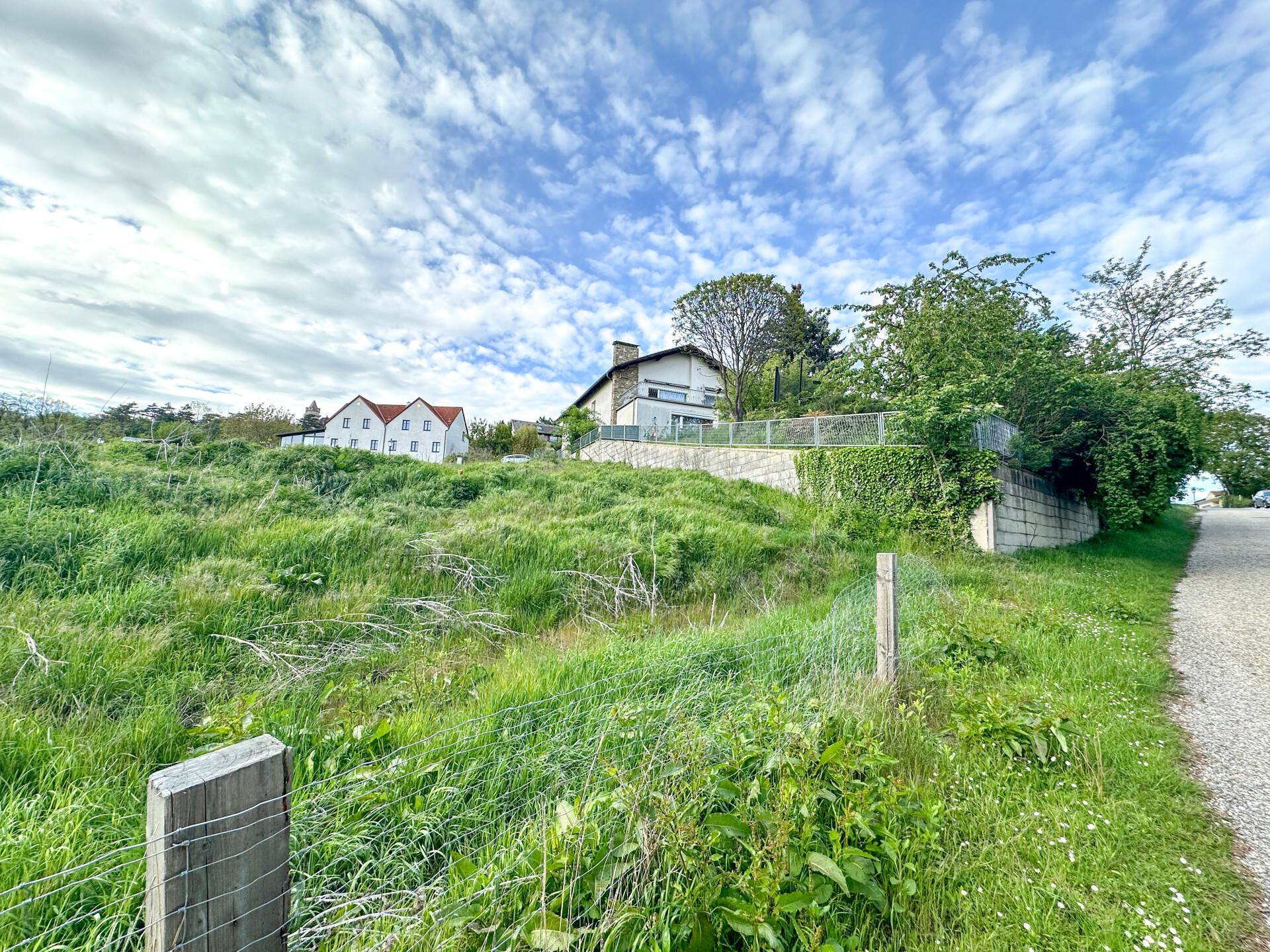 Hügeliges, grünes Gelände mit Drahtzaun und Wohnhäusern im Hintergrund unter einem teilweise bewölkten Himmel.
