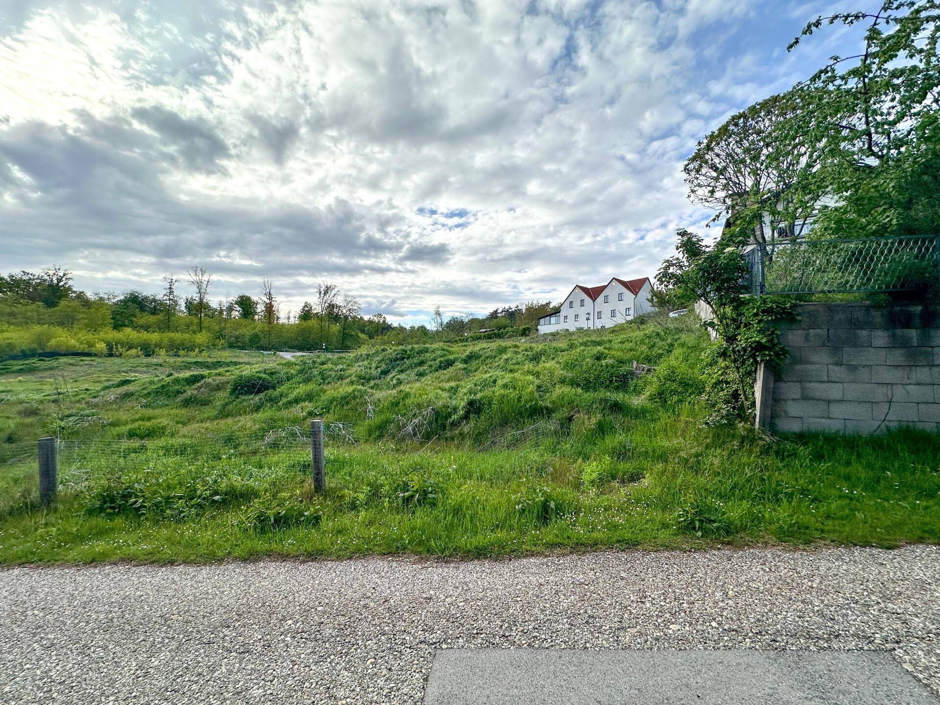 Grünes Hanggrundstück mit Schotterweg und einem traditionellen Haus mit rotem Dach in der Ferne.
