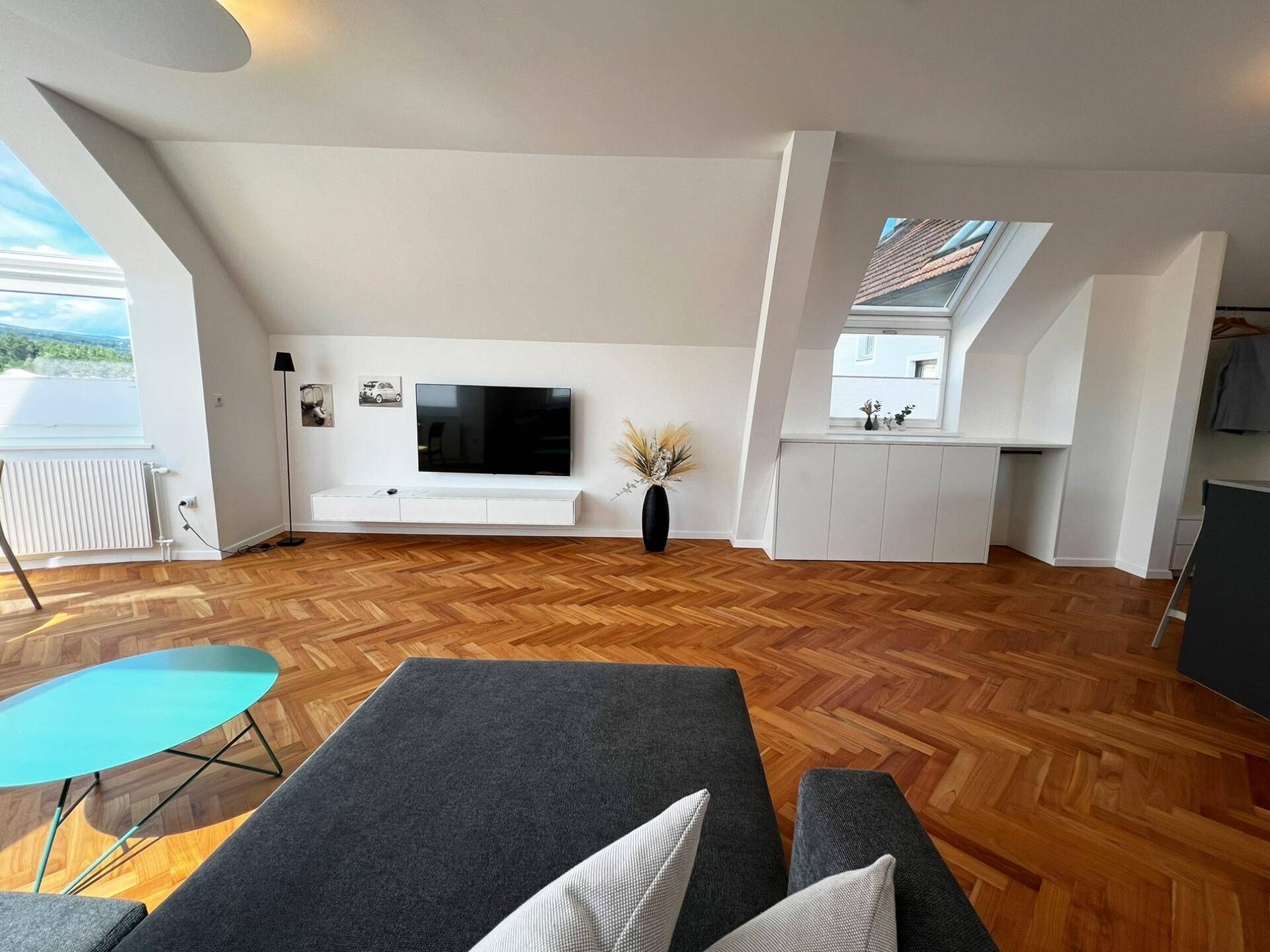 Geräumiger Wohnbereich mit Fischgrätparkett, Dachschrägen und einem großen Fernseher an der Wand.