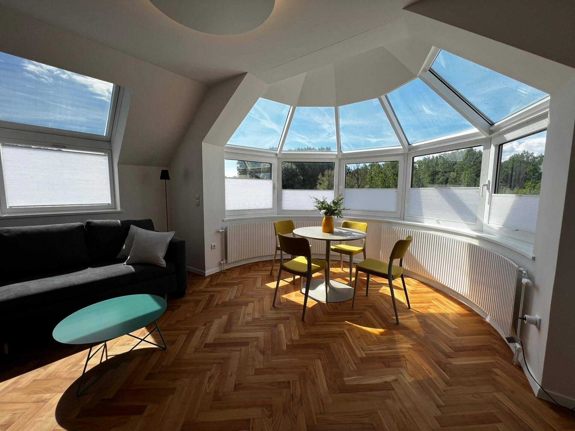 Heller Wohnbereich mit großen Panoramafenstern, Fischgrätparkett und moderner Couch unter Dachschrägen.