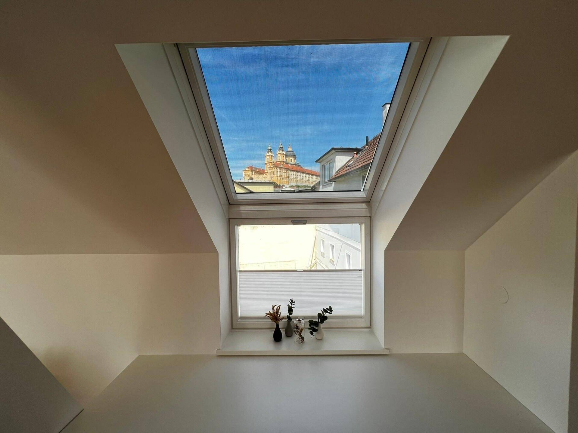 Dachfenster mit Blick auf historische Gebäude und den blauen Himmel, in einem hellen Innenraum.
