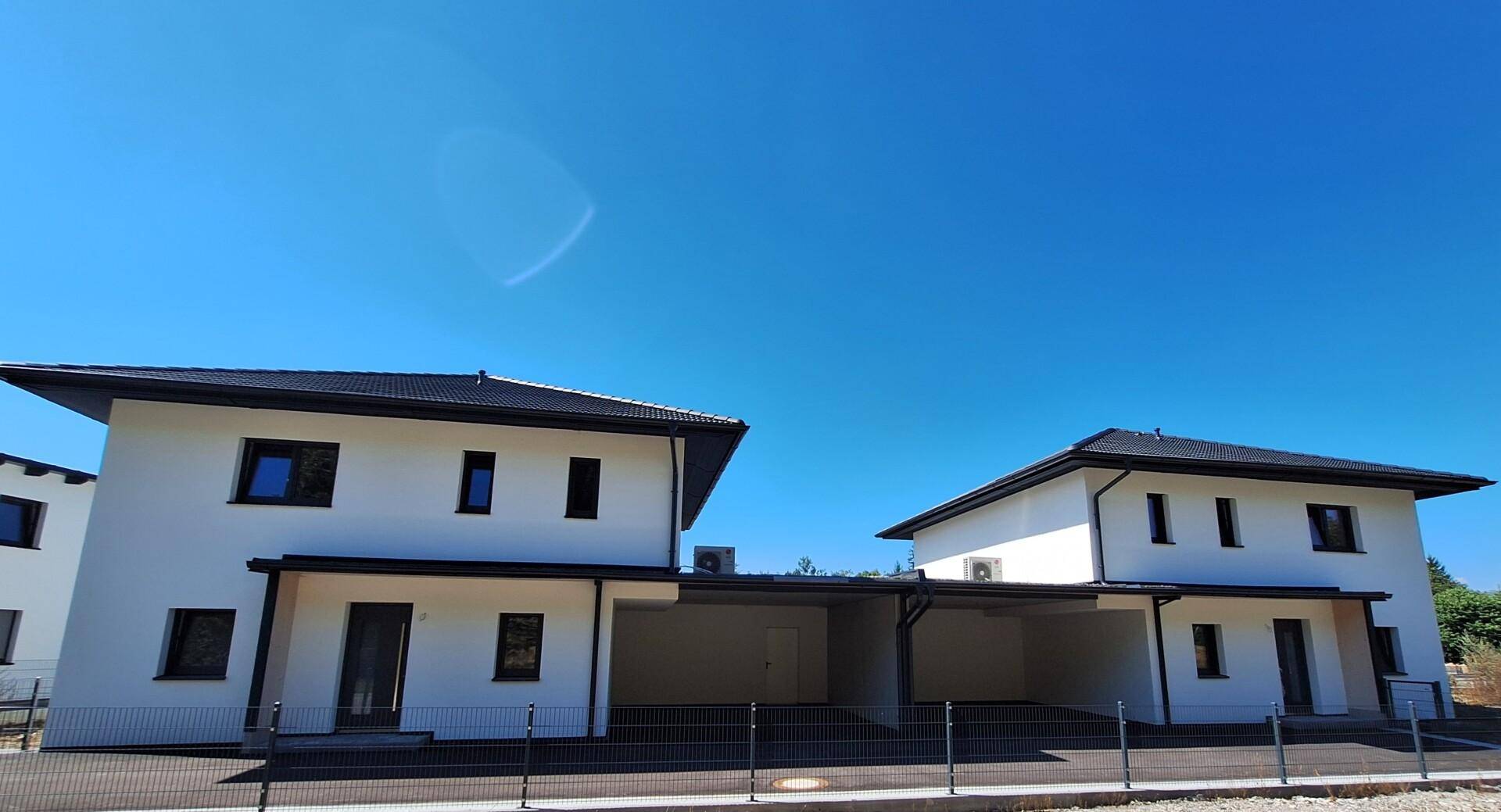 Zwei moderne Neubauhäuser mit weißer Fassade, dunklen Dächern und einem gemeinsamen Carport unter blauem Himmel.