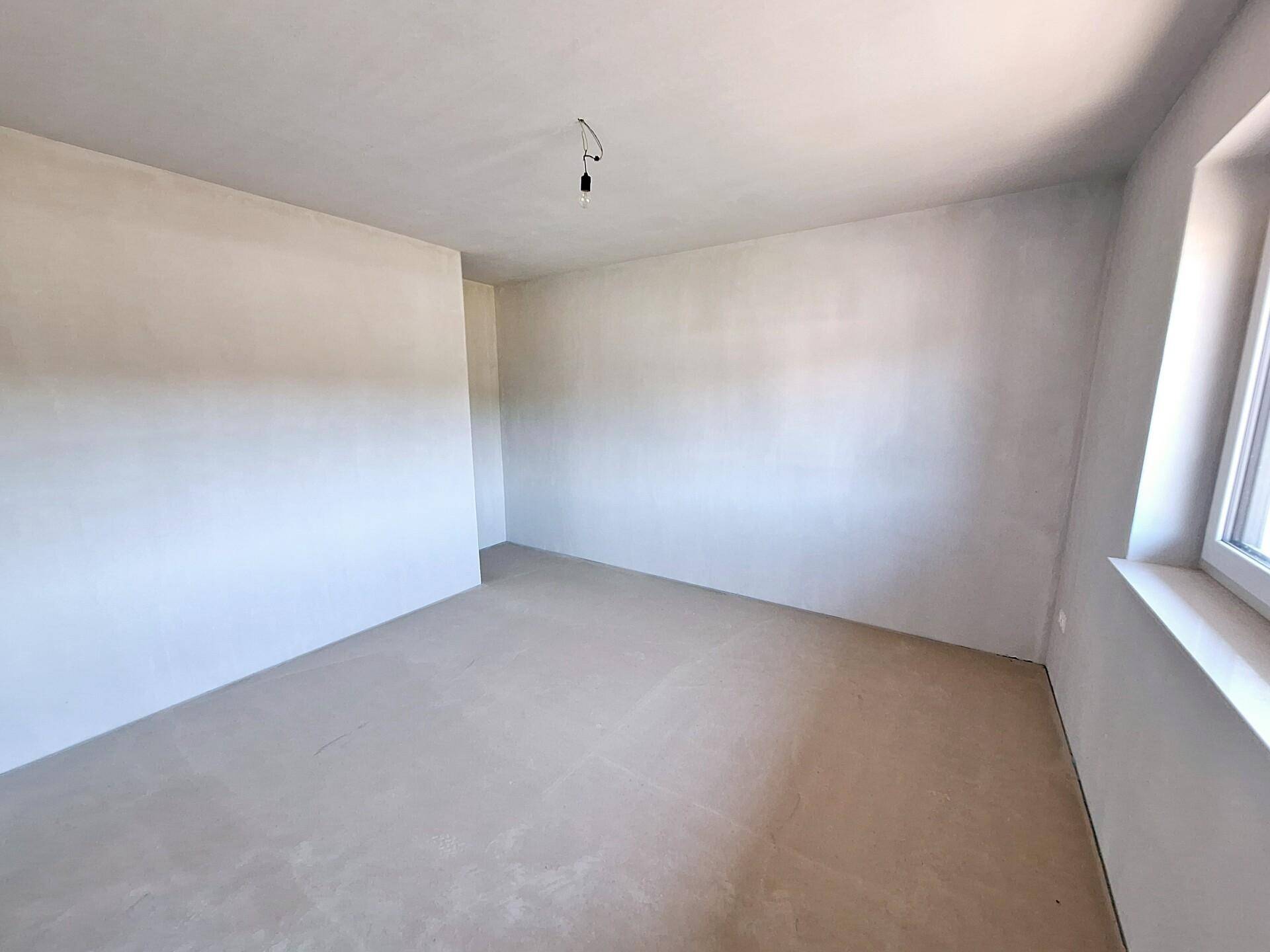 Leerer Raum im Rohbauzustand mit Fenster und hellem Betonboden, ideal für ein Schlafzimmer.