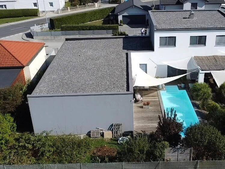 Luftaufnahme des modernen Gebäudes mit Pool, Holzterrasse und angrenzenden Grünflächen unter blauem Himmel.