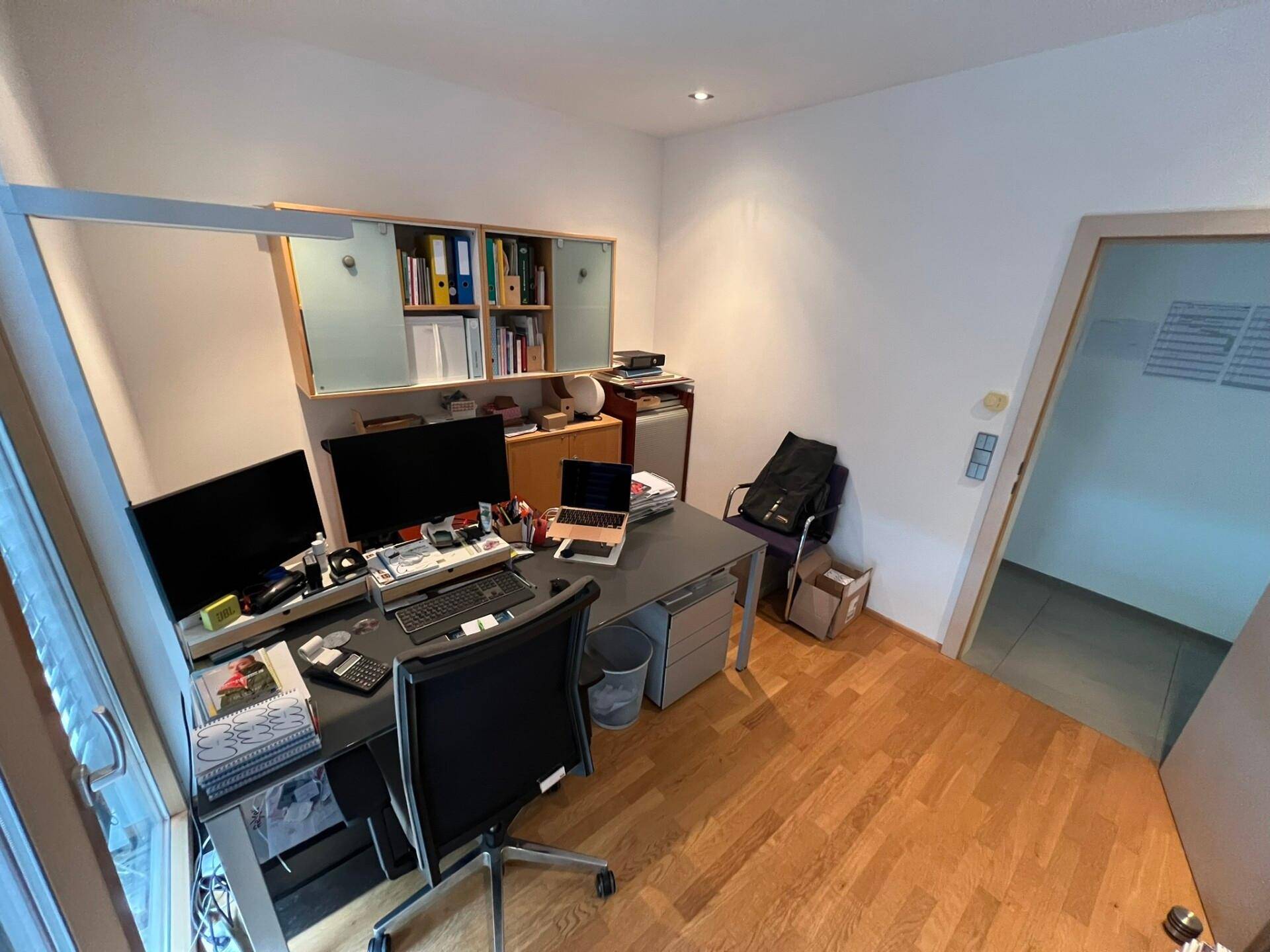 Funktionales Büro mit Holzparkett, einem Schreibtisch und einem Bücherregal, das durch eine Tür zugänglich ist.