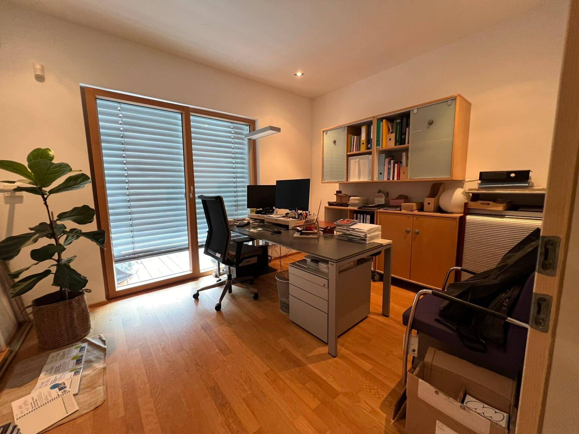 Modernes Büro mit Holzparkett, einem Schreibtisch und einem großen Fenster, das viel Tageslicht hereinlässt.
