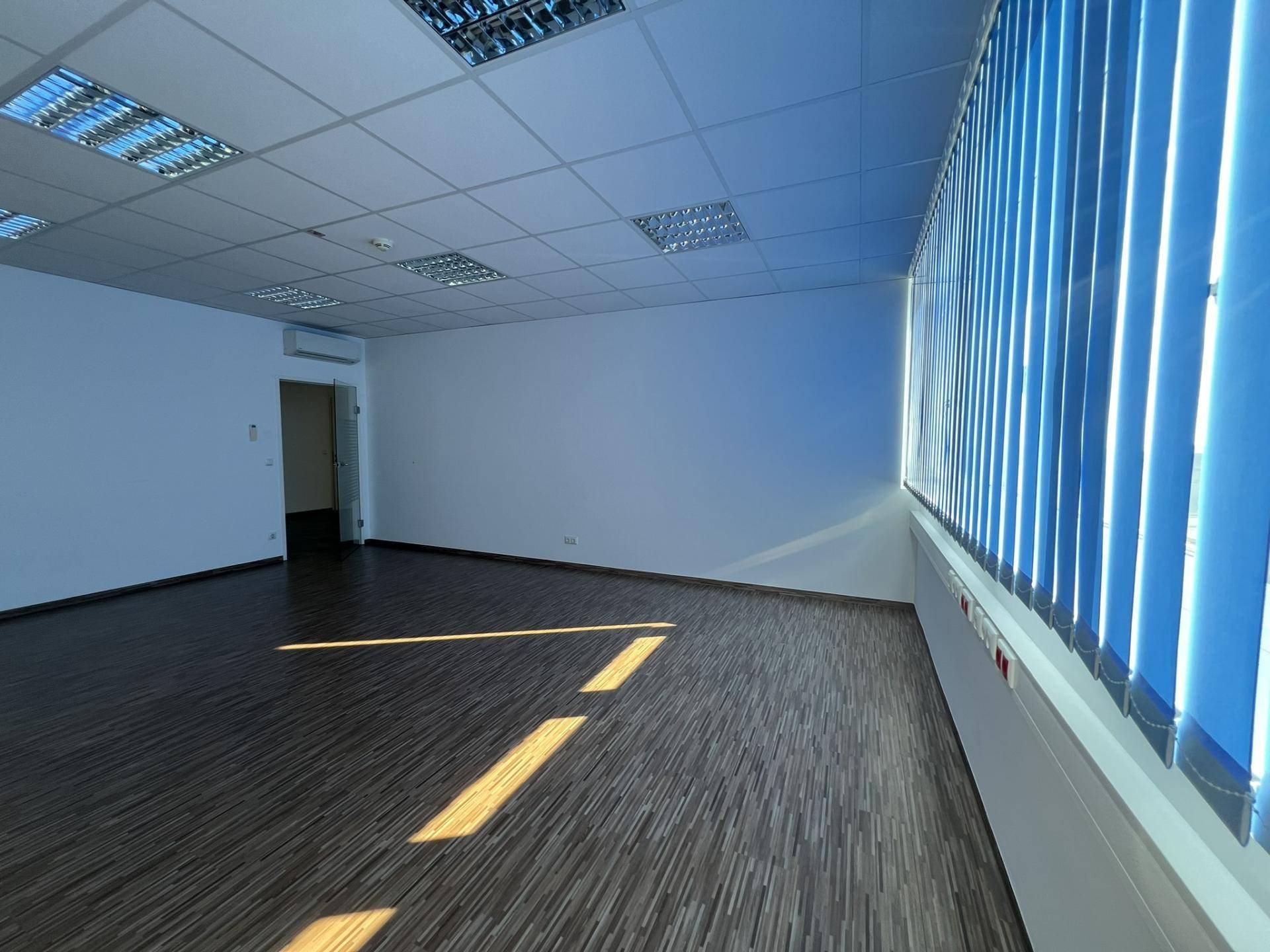 Geräumiger Büroraum mit dunklem Boden, weißen Wänden und einer langen Fensterfront mit blauen Jalousien, sehr hell.