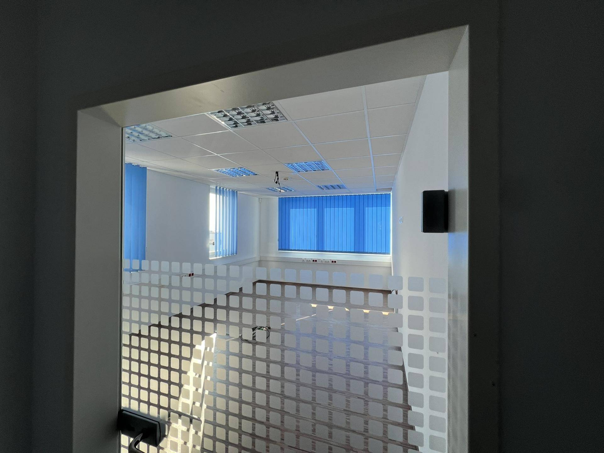 Blick durch eine Glastür in einen hellen Büroraum mit dunklem Boden und blauen Fensterjalousien, einladend und funktional.