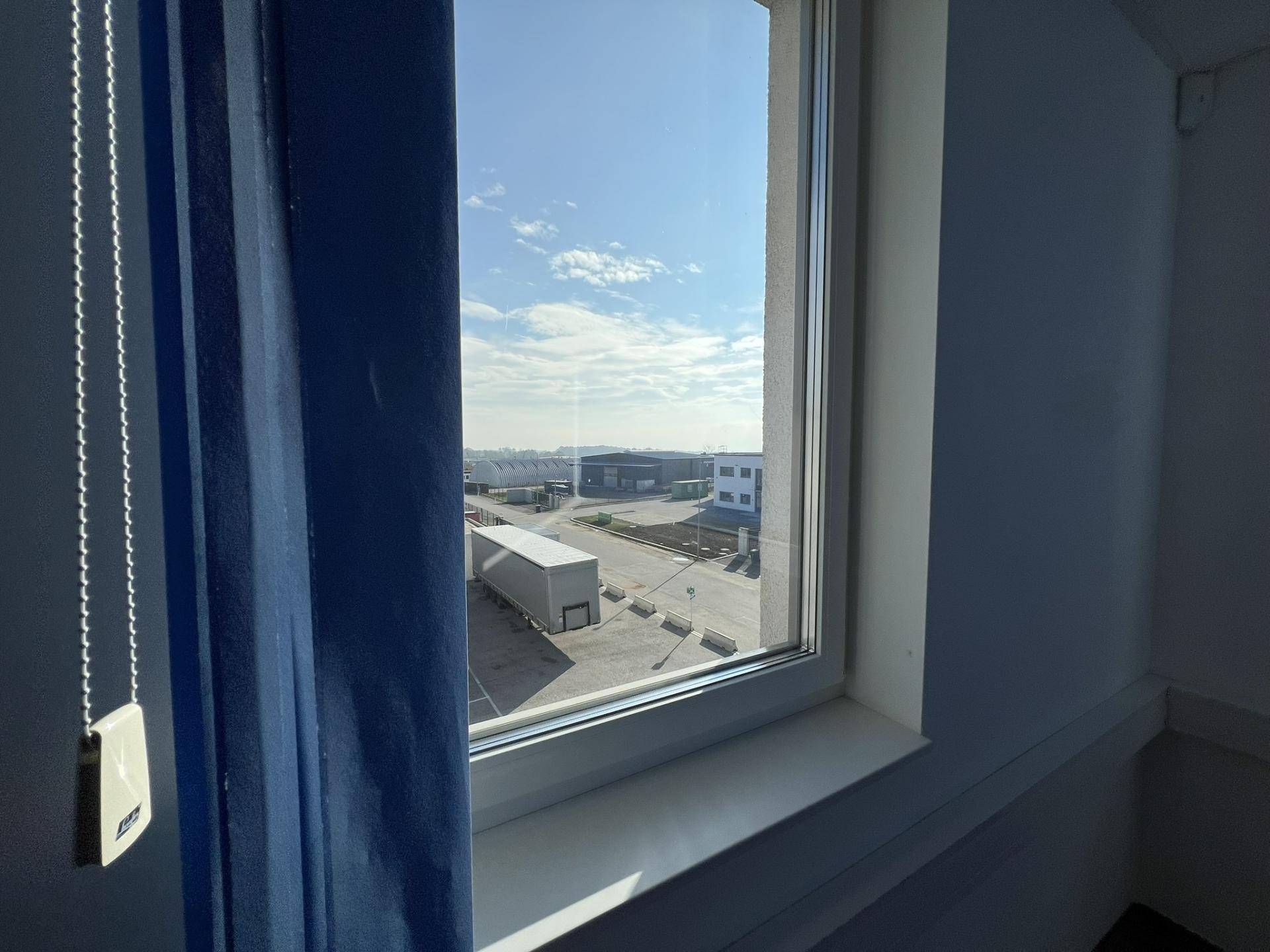 Ausblick aus einem Fenster auf das umliegende Gewerbegebiet mit Parkplätzen und einem LKW unter blauem Himmel.