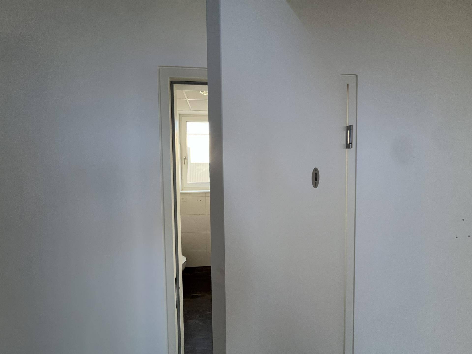 Blick durch eine offene Tür in einen hellen Raum mit Fenster, möglicherweise ein Badezimmer, sehr gepflegt.