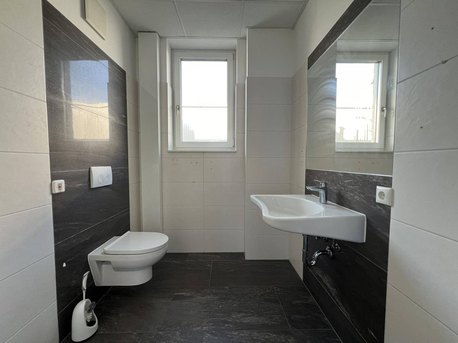 Modernes WC mit wandhängendem WC, Waschbecken, Fenster und dunklen sowie hellen Fliesen, viel Tageslicht.