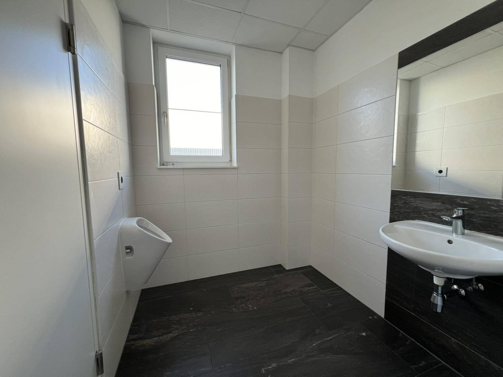 Modernes WC mit Urinal, Waschbecken, Fenster und dunklen Fliesen am Boden und an der Wand, sehr sauber.