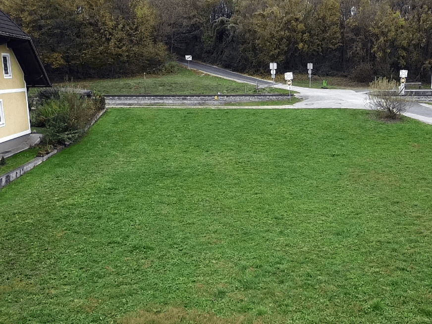 Blick auf eine große Rasenfläche neben einem Haus und einer Bahnlinie, umgeben von Bäumen.
