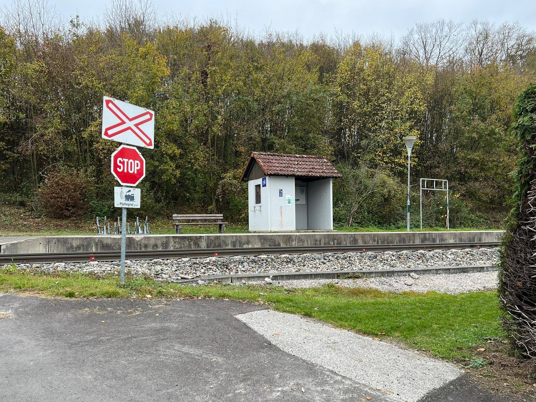 Kleiner Bahnsteig mit Wartehäuschen und Bahnübergangsschildern, umgeben von Bäumen und Büschen.