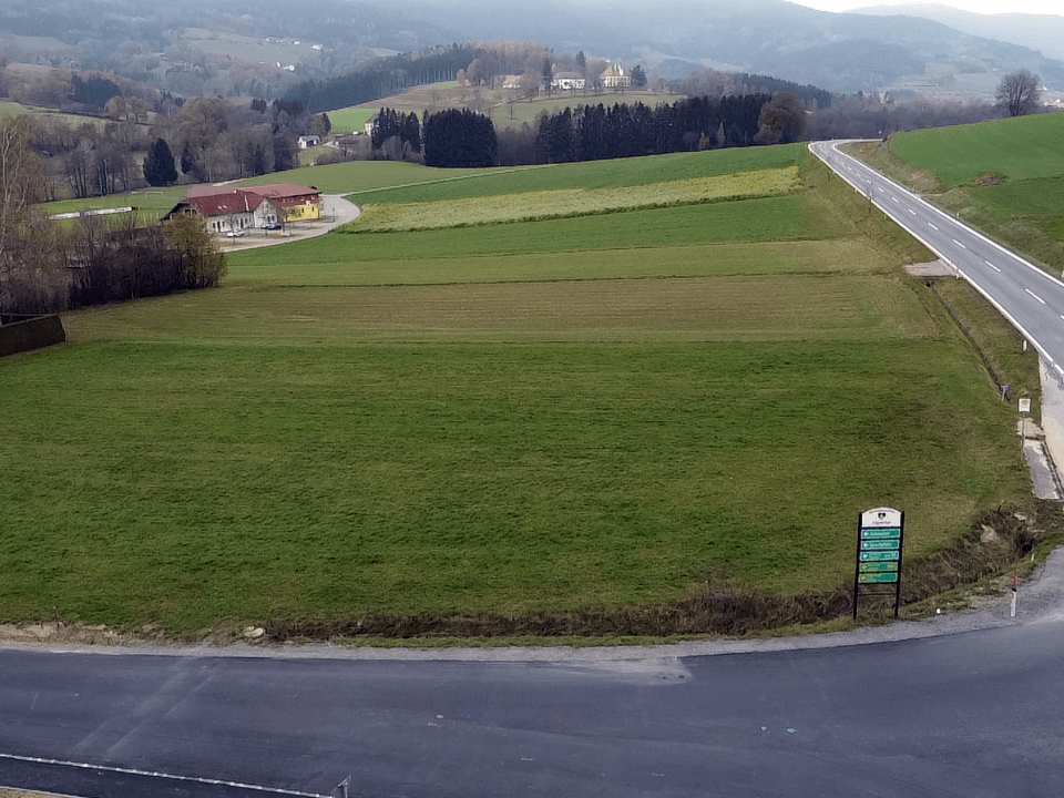 Weitläufige Landschaftsaufnahme mit grünen Feldern, einer kurvigen Straße und vereinzelten Gebäuden vor einer bergigen Kulisse.