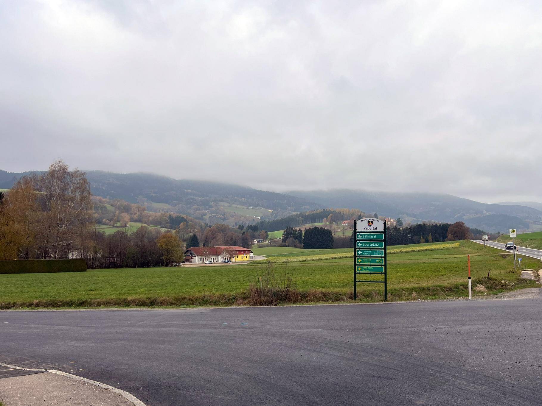 Panoramablick auf eine ländliche Straße, umgeben von grünen Wiesen und bewaldeten Hügeln unter einem bewölkten Himmel.