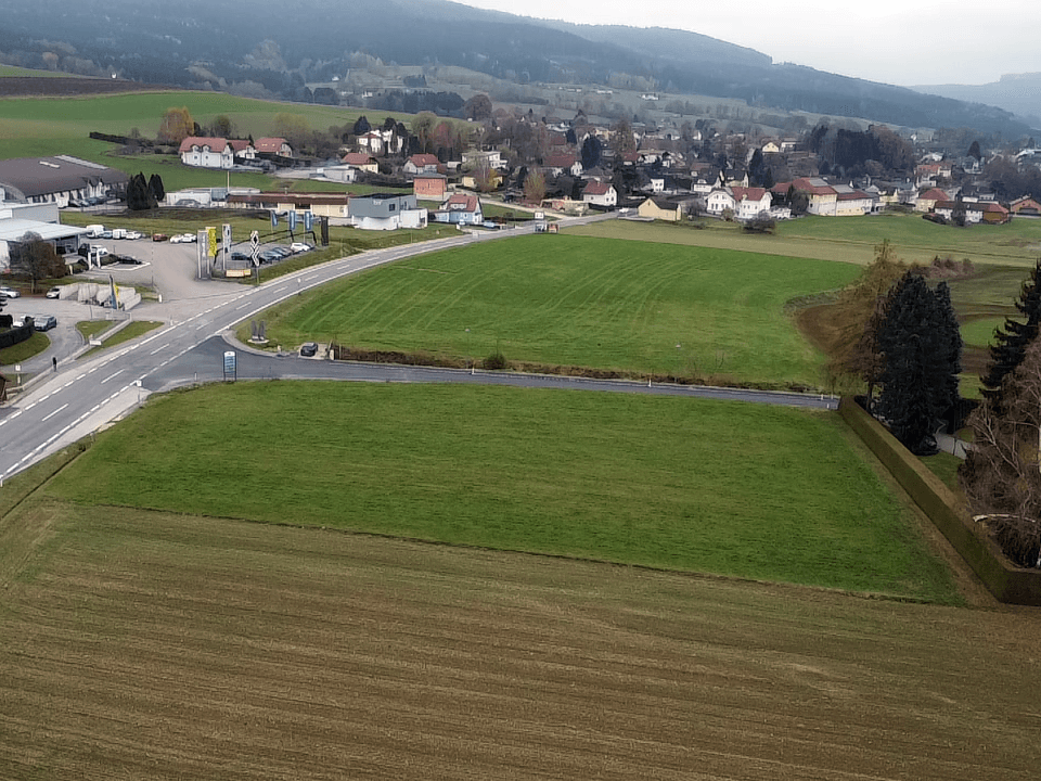 Luftaufnahme eines grünen Baugrundstücks neben einer Straße, umgeben von Feldern und einem Dorf in der Ferne.