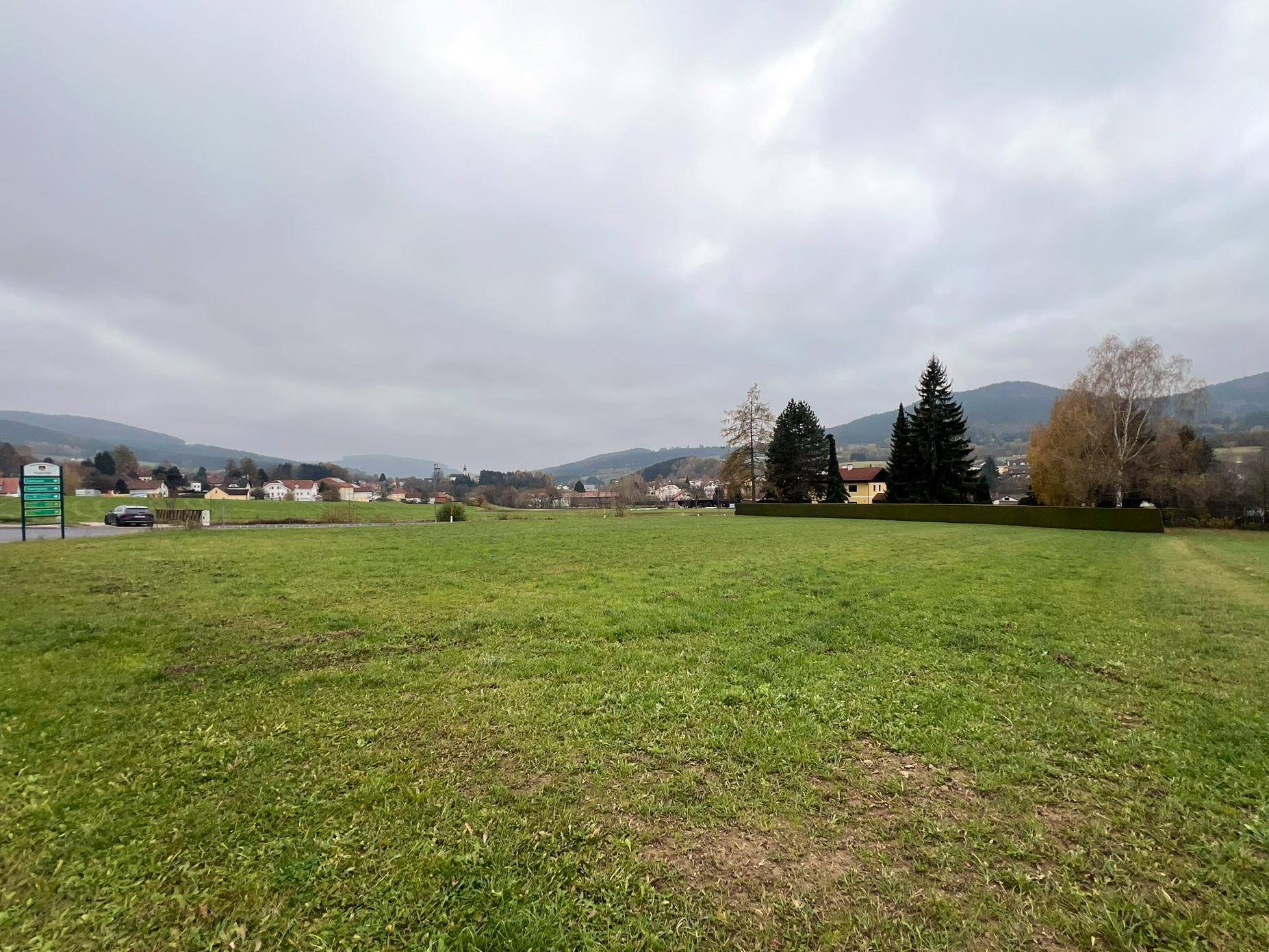 Weitläufiges grünes Grundstück mit Blick auf ein entferntes Dorf und bewaldete Hügel unter einem grauen Himmel.