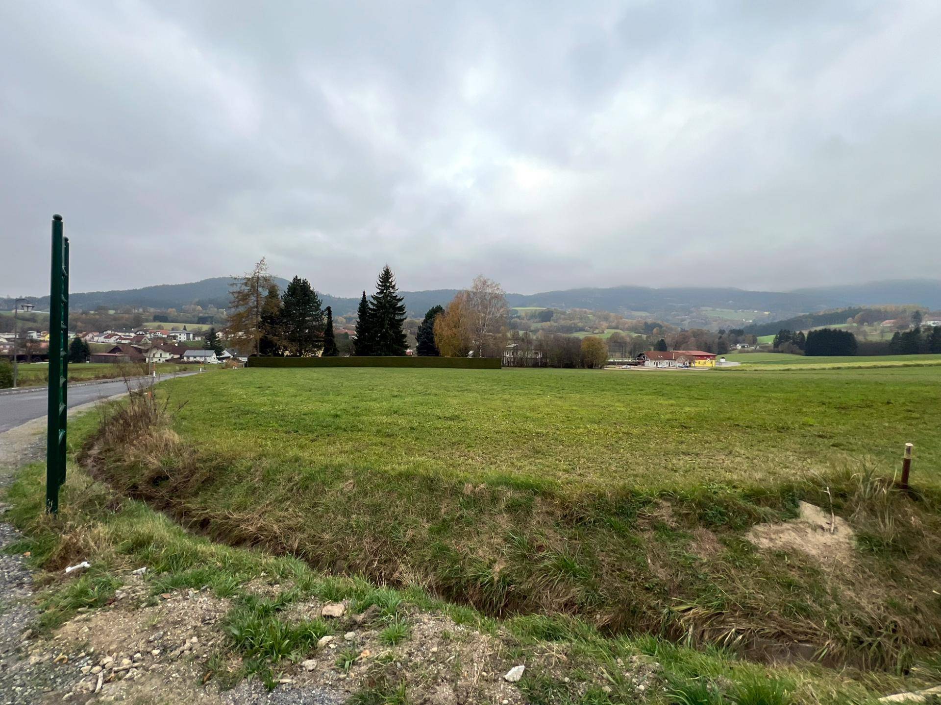 Grünes Grundstück mit Blick auf eine ländliche Straße und bewaldete Hügel unter einem bewölkten Himmel.