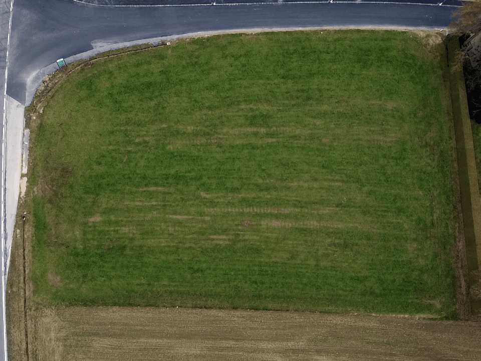 Senkrechte Luftaufnahme eines rechteckigen grünen Baugrundstücks, angrenzend an eine Straße und weitere Felder.