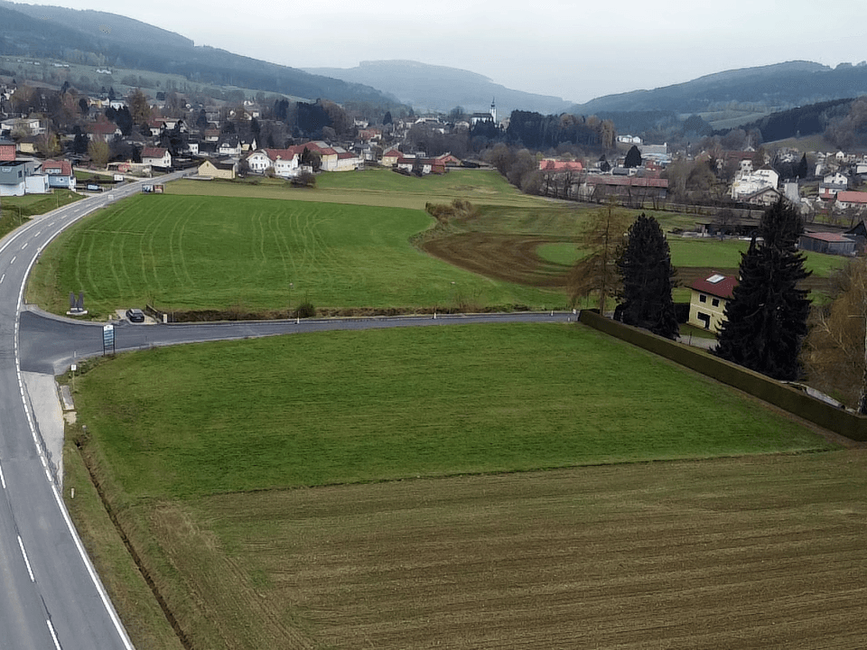 Luftaufnahme eines grünen Baugrundstücks mit gelber Umrandung, umgeben von Feldern und einer Straße, mit einem Dorf im Hintergrund.