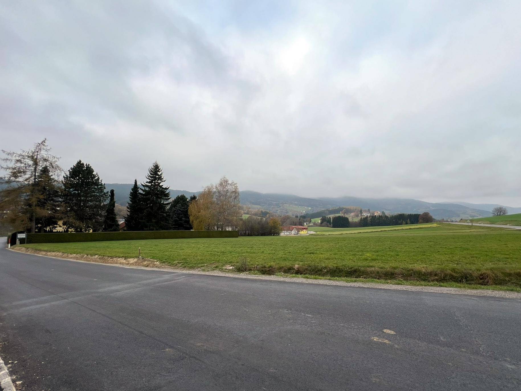 Weitläufige grüne Landschaft mit einer Straße im Vordergrund und bewaldeten Hügeln in der Ferne unter einem grauen Himmel.
