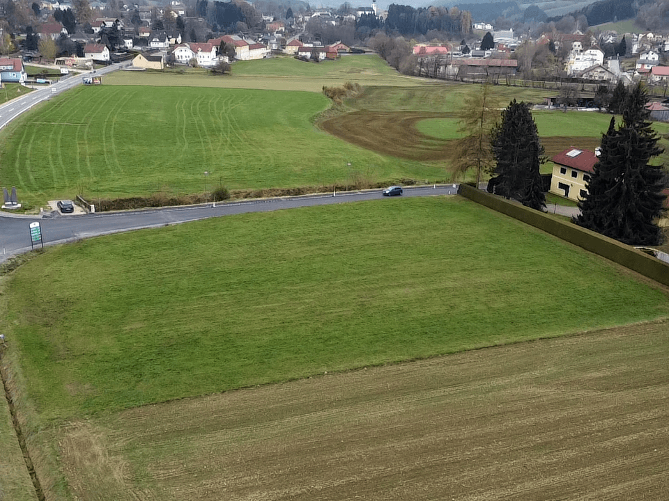 Luftaufnahme eines grünen Baugrundstücks mit gelber Umrandung, umgeben von Feldern und einer Straße, mit einem Dorf im Hintergrund.