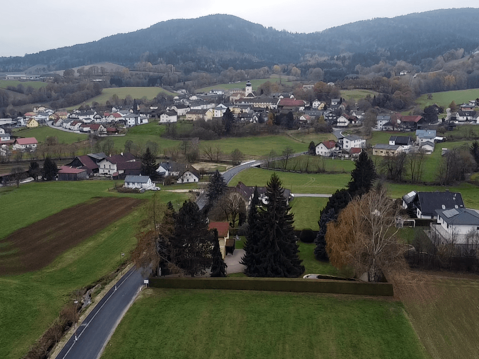 Panoramablick aus der Vogelperspektive auf ein malerisches Dorf, eingebettet in eine grüne Hügellandschaft mit Wäldern.