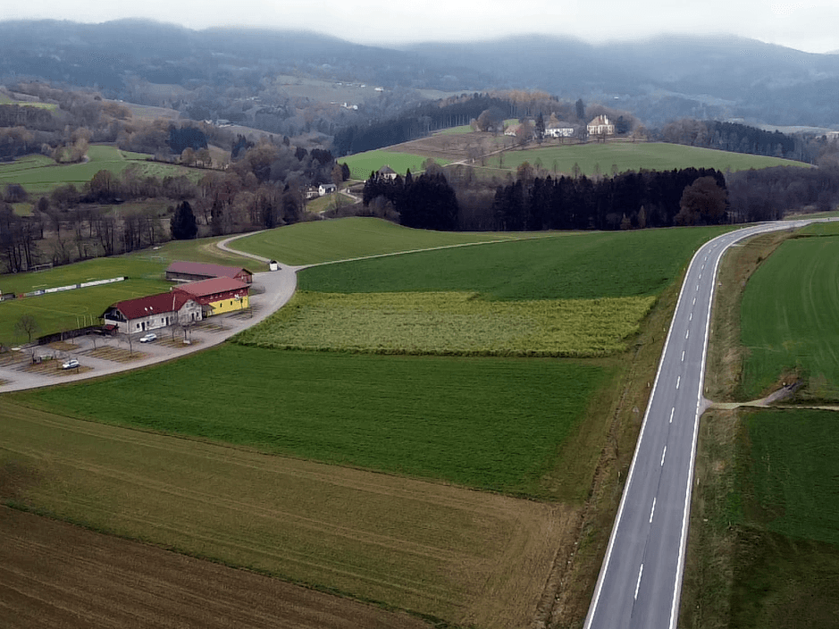 Weitläufige Luftaufnahme einer ländlichen Straße, umgeben von grünen Feldern und bewaldeten Hügeln unter einem bewölkten Himmel.