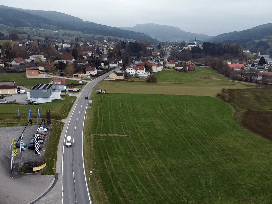 Luftaufnahme einer Landstraße, die sich durch grüne Felder schlängelt, mit einem Dorf und Hügeln im Hintergrund.