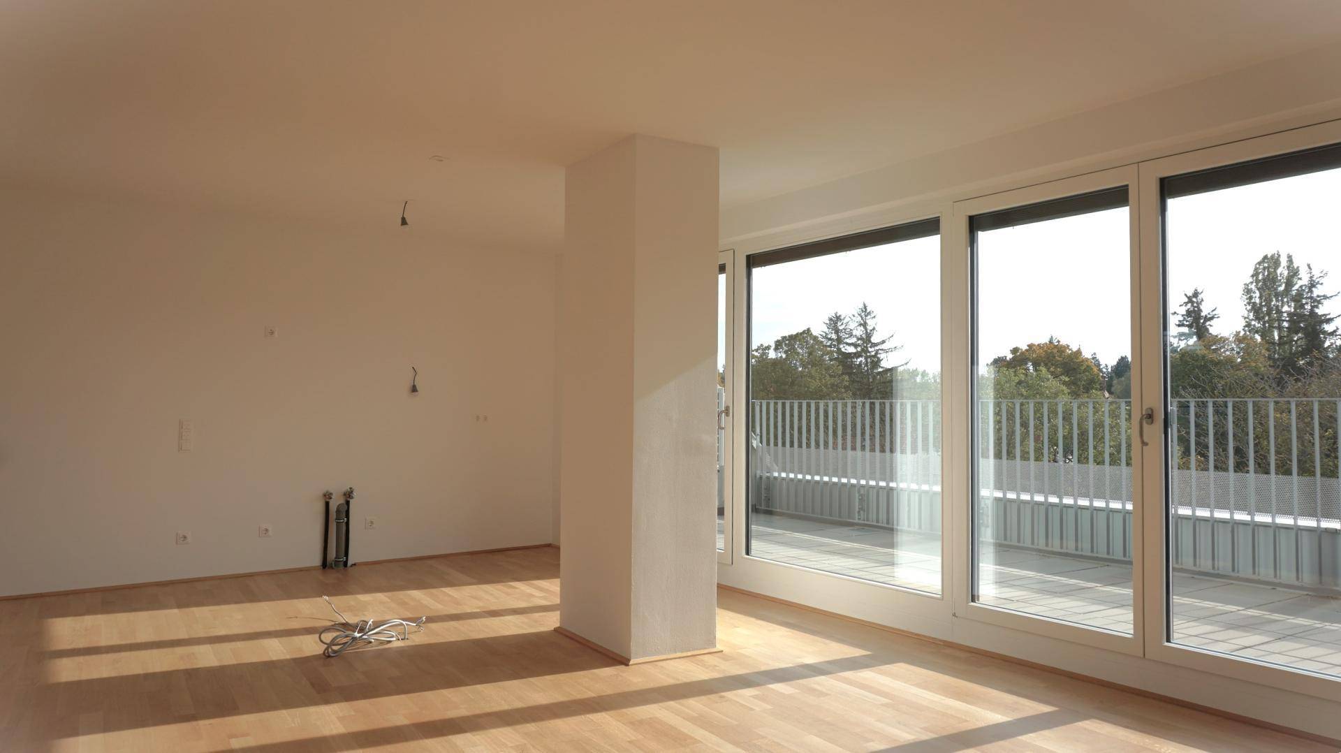 Leerer, lichtdurchfluteter Raum mit Holzboden und großen Fenstern, die Zugang zu einer Terrasse bieten.