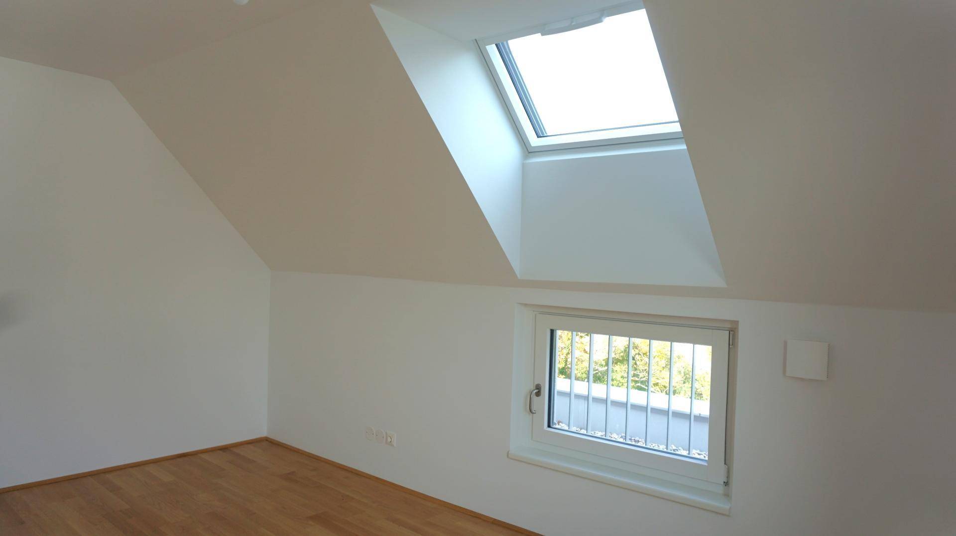 Leerer Raum im Dachgeschoss mit schrägen Wänden, Holzboden und einem großen Dachfenster.