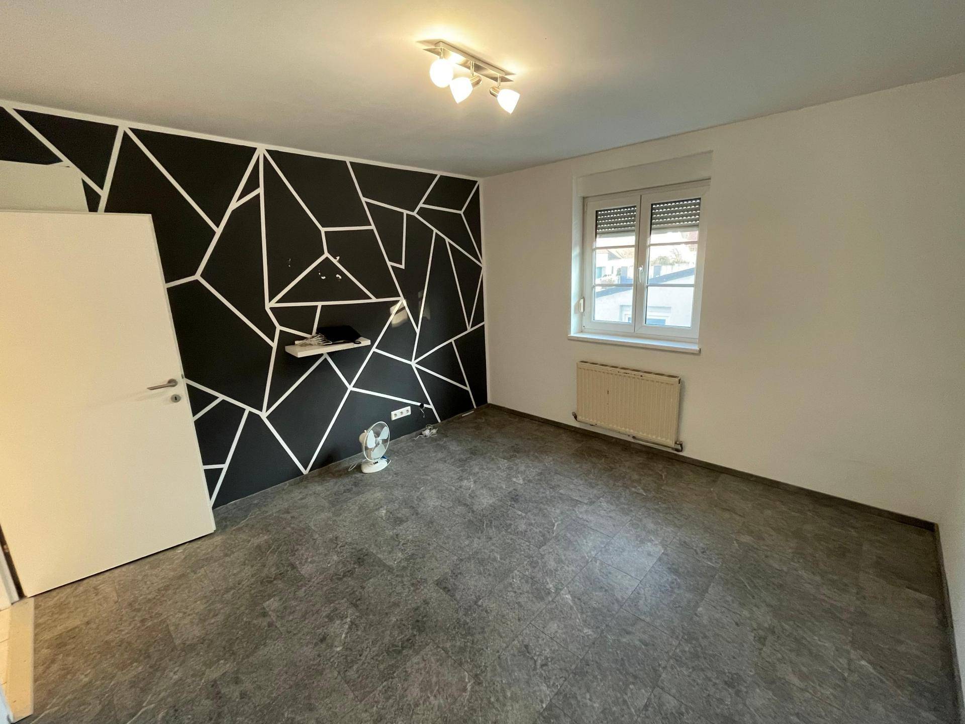 Schlafzimmer mit modernem geometrischem Wanddesign, dunklem Boden und Fenster mit Rollladen.