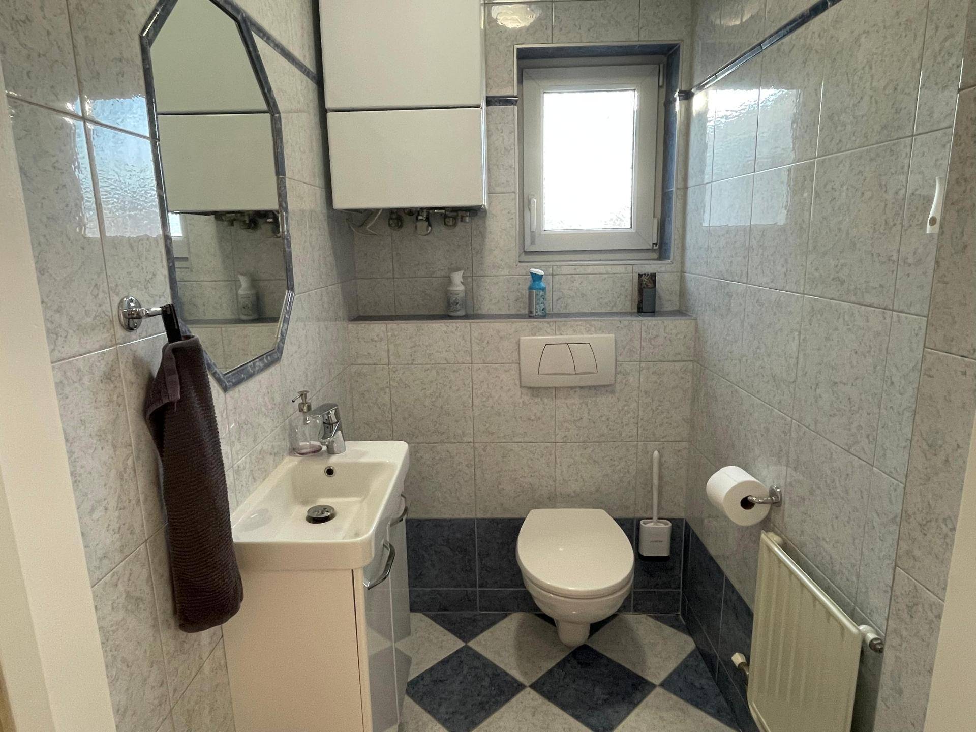 Kleines Gäste-WC mit WC, Waschbecken, Fenster und gemusterten Fliesen im 80er-Jahre-Stil.