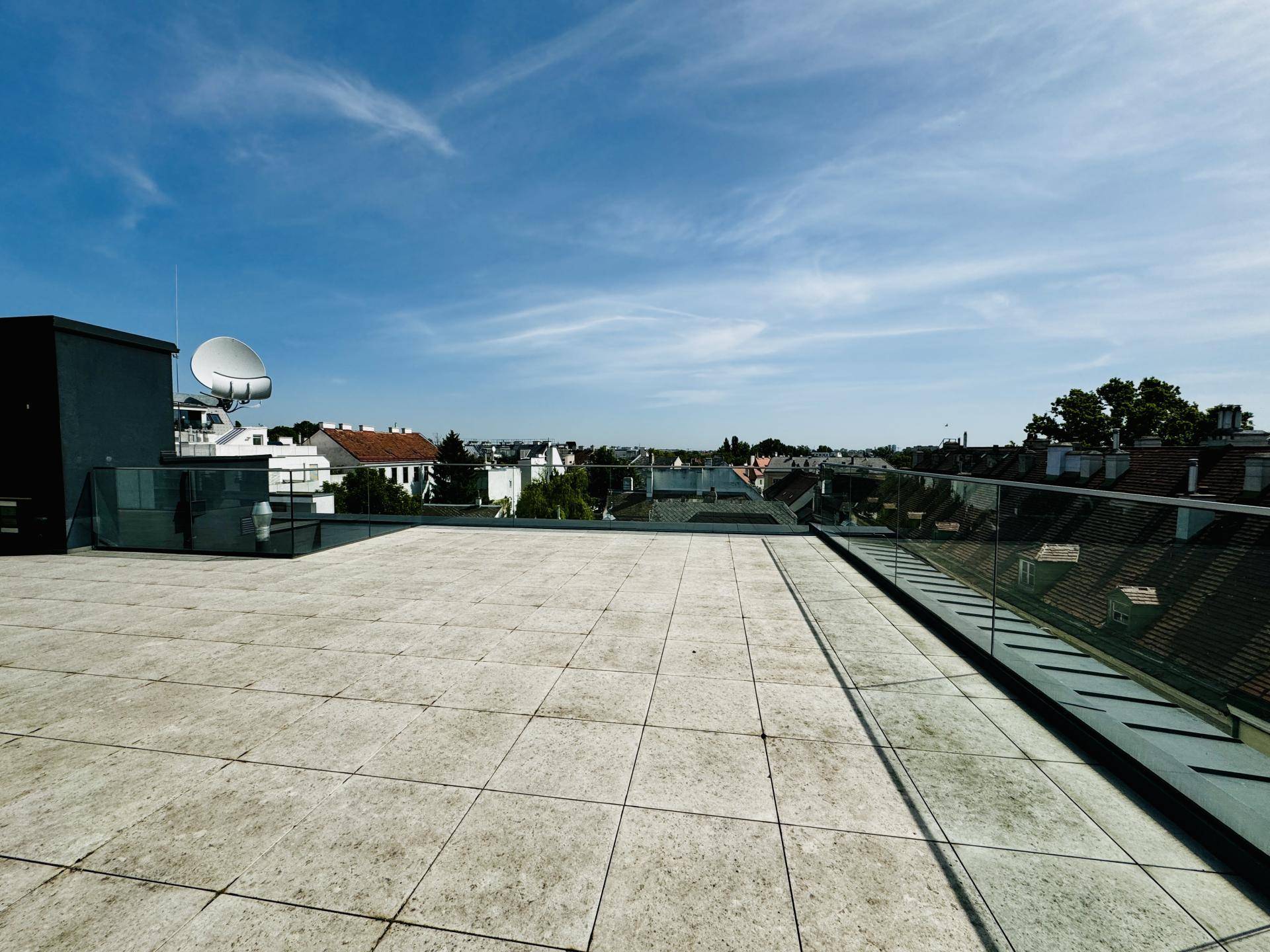 Geräumige Dachterrasse mit Fliesenboden und Glasgeländer, bietet weite Ausblicke unter blauem Himmel.