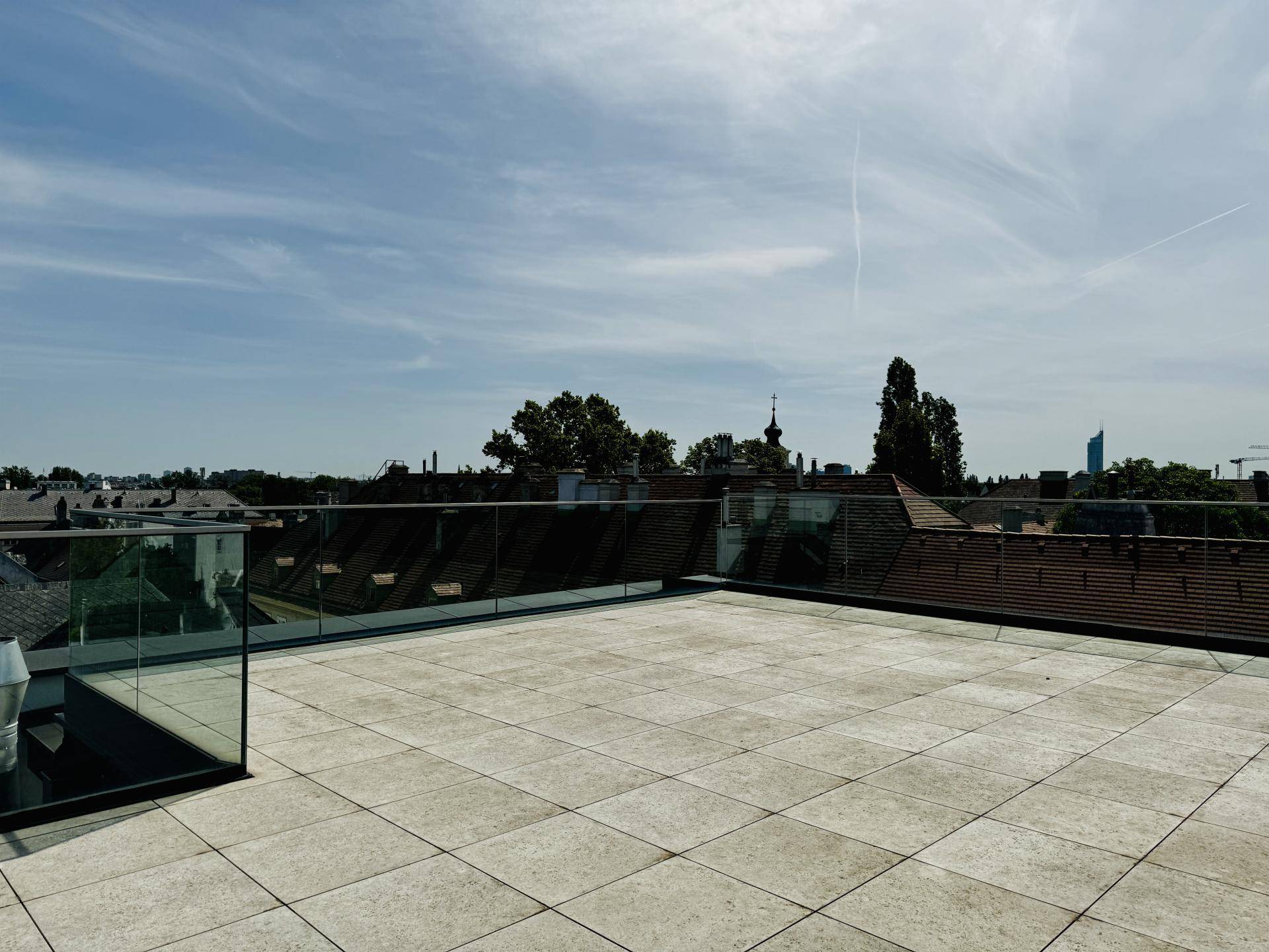 Geräumige Dachterrasse mit Fliesenboden und Glasgeländer, bietet weite Ausblicke unter blauem Himmel.