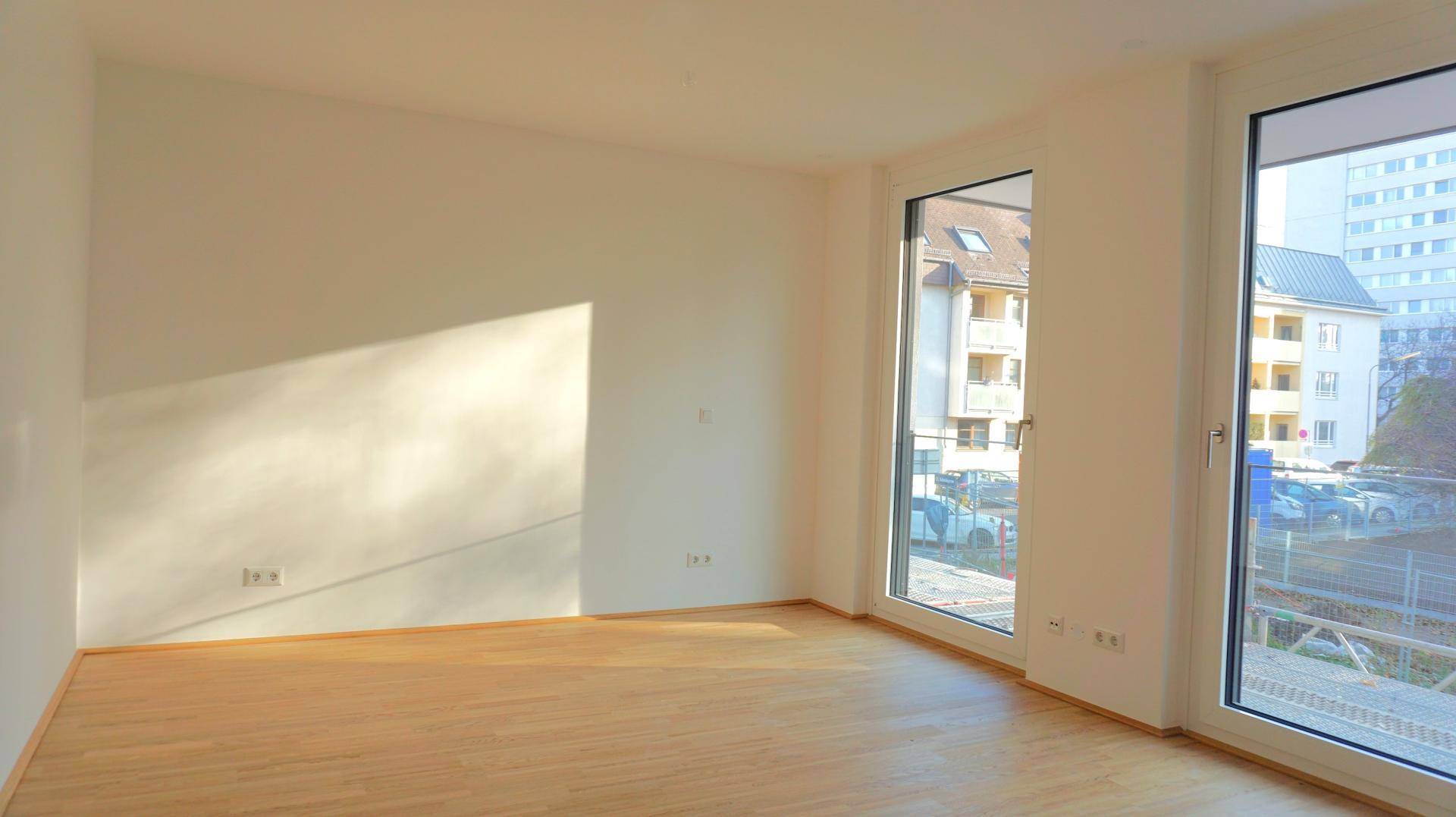 Helles, leeres Zimmer mit großen Fenstern und hellem Parkettboden, sonnendurchflutet.