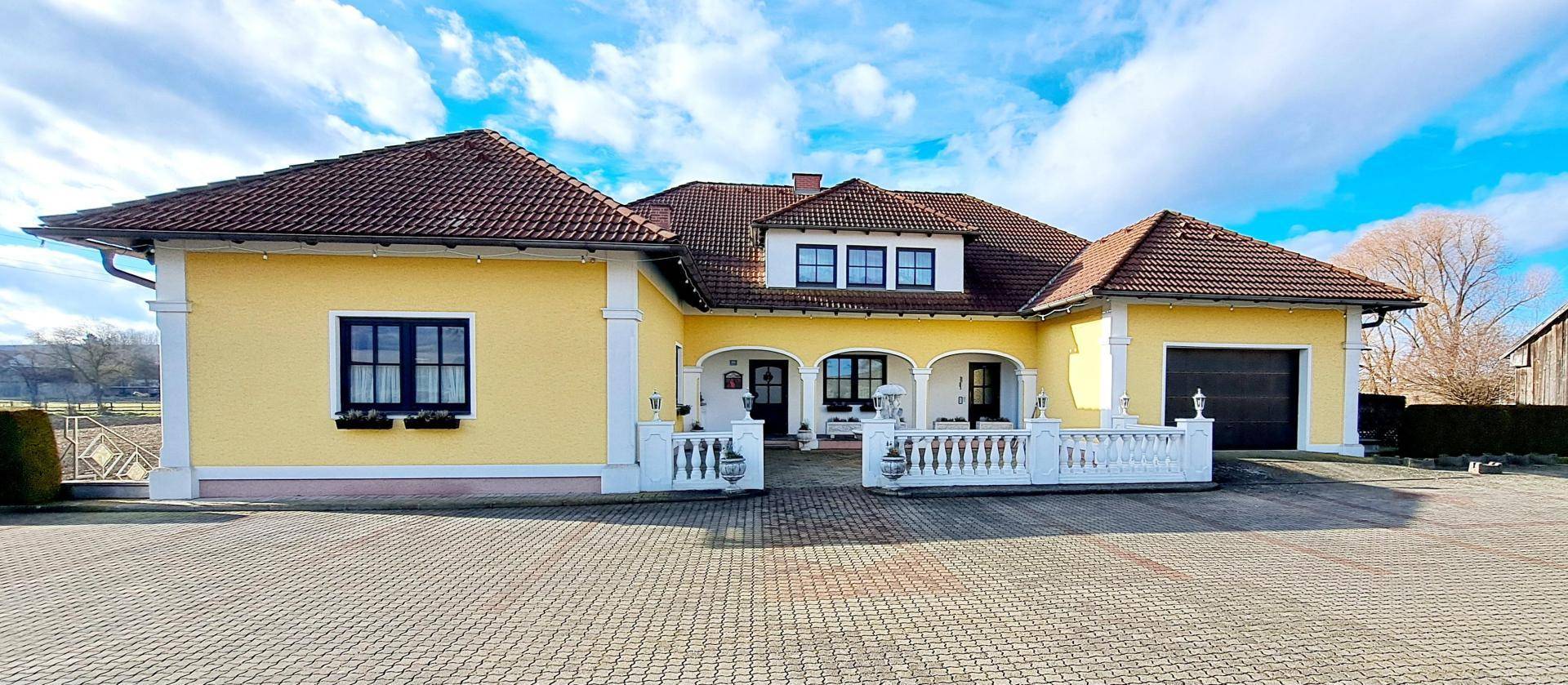 Charmantes Landhaus mit gelber Fassade, weißen Balustraden und gepflastertem Vorplatz.