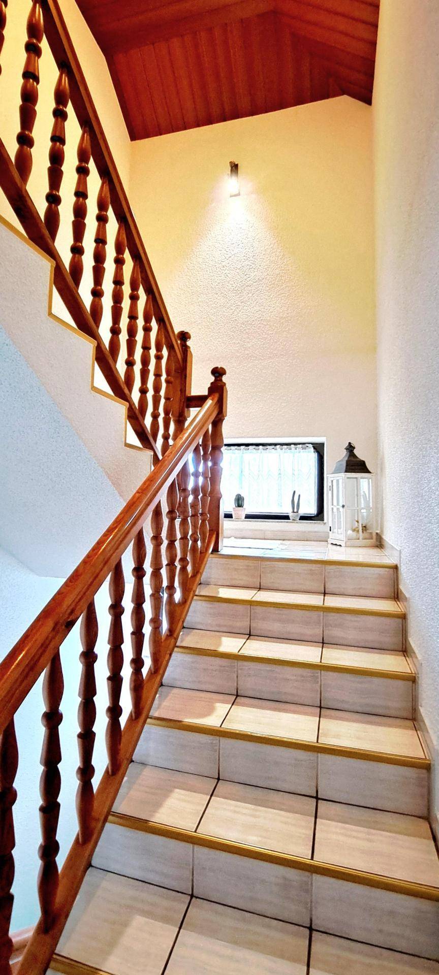 Helles Treppenhaus mit Holzgeländer, Fliesenstufen und Fenster für natürliches Licht.